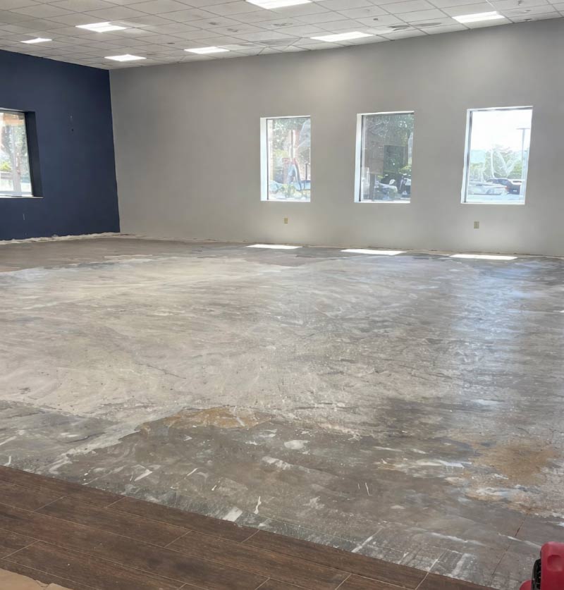 A large commercial interior space during the initial stages of a renovation, showcasing a bare and unfinished concrete floor with visible patches of adhesive and leveling compound. The room features high ceilings with a white drop-tile grid and recessed square LED lighting. One wall is painted a deep navy blue, while the adjacent wall is a light gray with three vertical rectangular windows letting in natural light. In the foreground, a small section of dark wood-look plank flooring has been laid, contrasting with the raw subfloor.