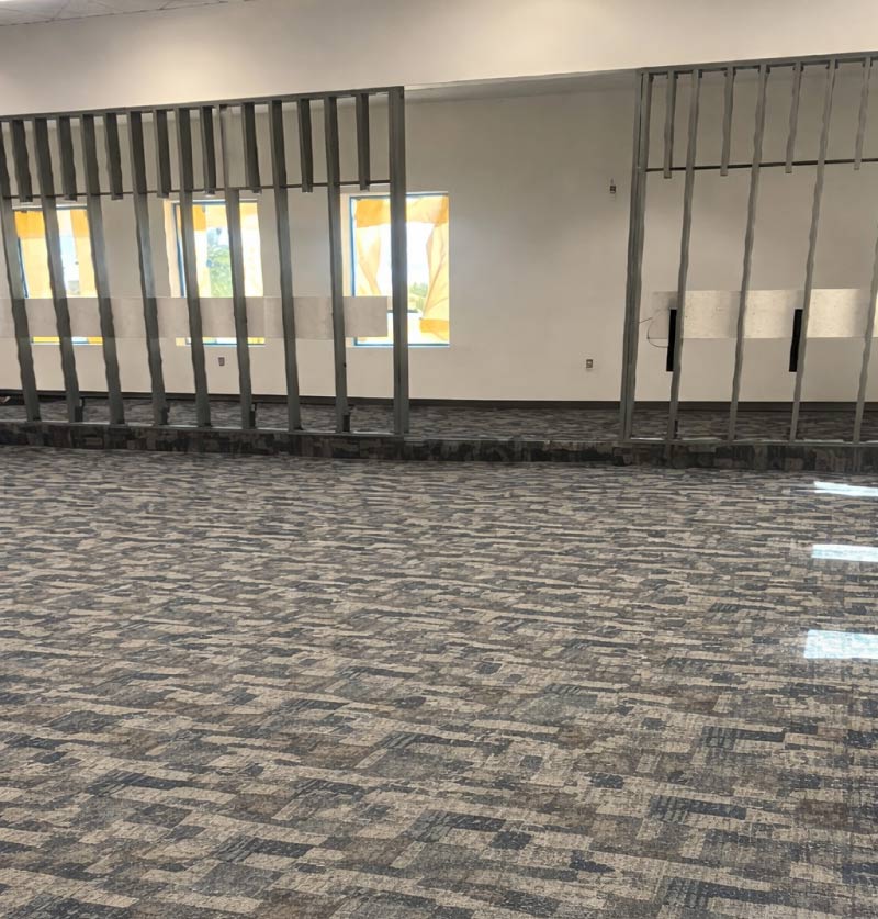 The completed interior of a modern commercial office space featuring newly installed patterned carpet tiles in shades of gray, blue, and tan. The room is partitioned by open metal stud framing, indicating the layout of future office walls. In the background, three large windows are visible, partially covered with yellow protective film, allowing soft light to filter onto the textured carpet. The walls are finished in a clean, neutral white, and the overhead lighting from the previous stage illuminates the professional and updated environment.