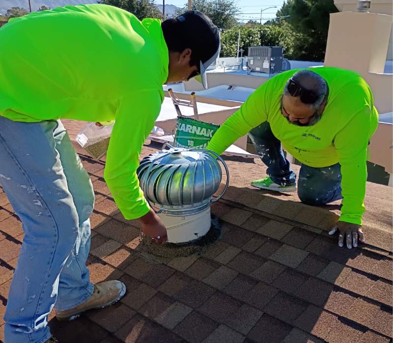 Guide to Roof Damage: Two JM Roofing and Construction team members working together to repair a silver turbine roof vent on a sloped shingle roof. One technician in a neon safety shirt applies sealant around the base of the vent while the other assists with professional-grade Karnak roofing cement. This detailed repair process prevents water intrusion and ensures the residential roofing system remains properly ventilated and leak-free.