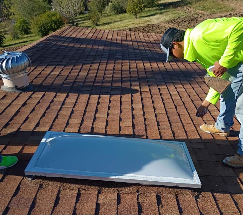 A professional roofer from JM Roofing and Construction wearing a high-visibility safety shirt performing a specialized skylight seal repair on a residential shingle roof. The technician is carefully applying granules from a container to a freshly sealed skylight frame to ensure a UV-resistant and watertight finish that matches the brown architectural shingles. A silver turbine roof vent is visible in the background, showing a comprehensive approach to roof ventilation and maintenance.