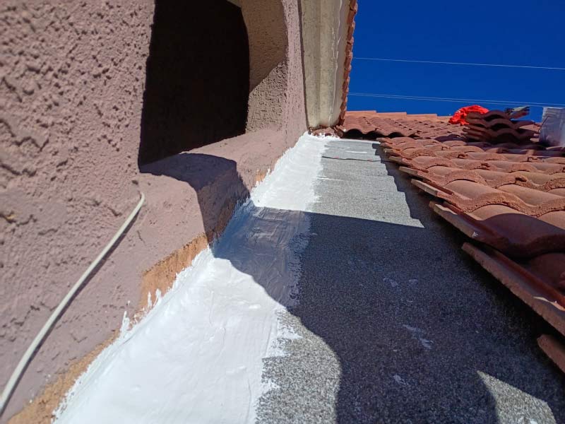Close-up view of a professional roof coating application on a tile roofing system where the roof meets a stucco wall. A thick, white waterproof sealant has been meticulously applied along the flashing and transition area to prevent leaks in high-risk valley locations. The contrast between the red clay tiles and the fresh white protective coating highlights the precision of the maintenance work performed by JM Roofing and Construction.