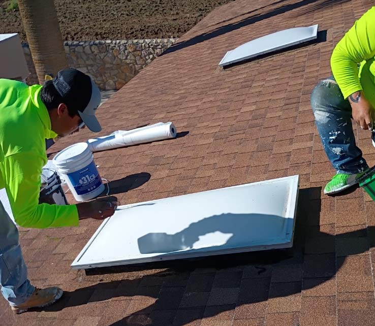 Two JM Roofing and Construction professionals in high-visibility safety gear meticulously apply specialized sealant to a residential skylight on a brown shingle asphalt roof in El Paso. One technician uses a precision tool to ensure a watertight bond, demonstrating the company’s commitment to high-quality standards and detailed maintenance.
