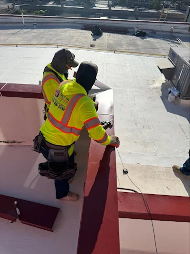 Professional JM Roofing contractors in high-visibility safety vests working on the red perimeter metal flashing of a large commercial flat roofing project.