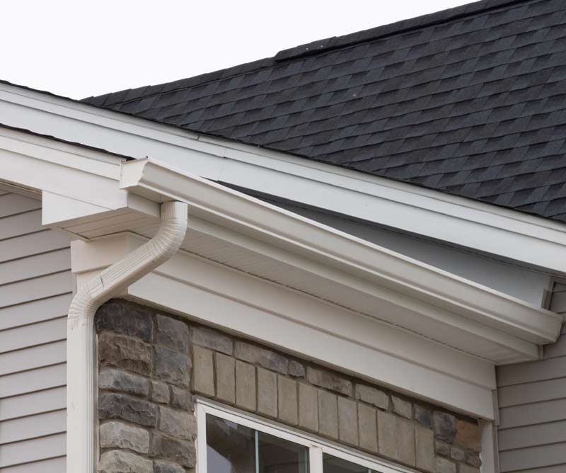 Dark architectural asphalt shingle roof featuring clean white K-style gutters and a downspout against vinyl and stone siding.